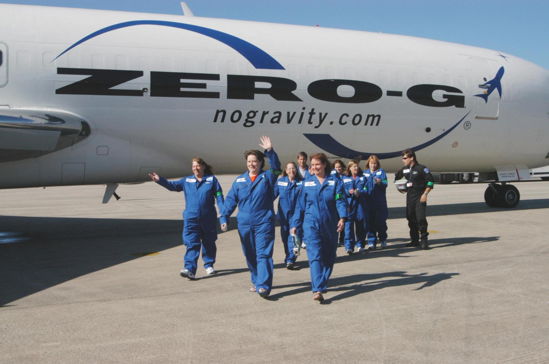 Image description: A group of people, dressed in blue jumpsuits, walk toward the camera away from a Zero-G plane parked on an airport runway. End of alt text. 