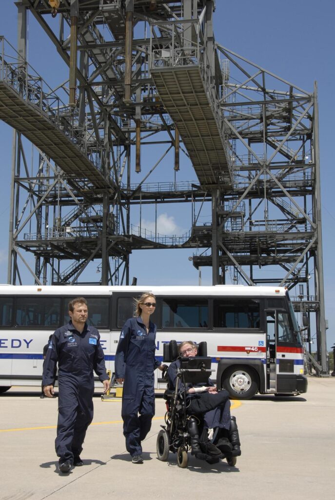Image description: In the foreground, two Zero-G workers accompany Stephen Hawking; behind is a stationary bus and a metal framework. End of alt text.