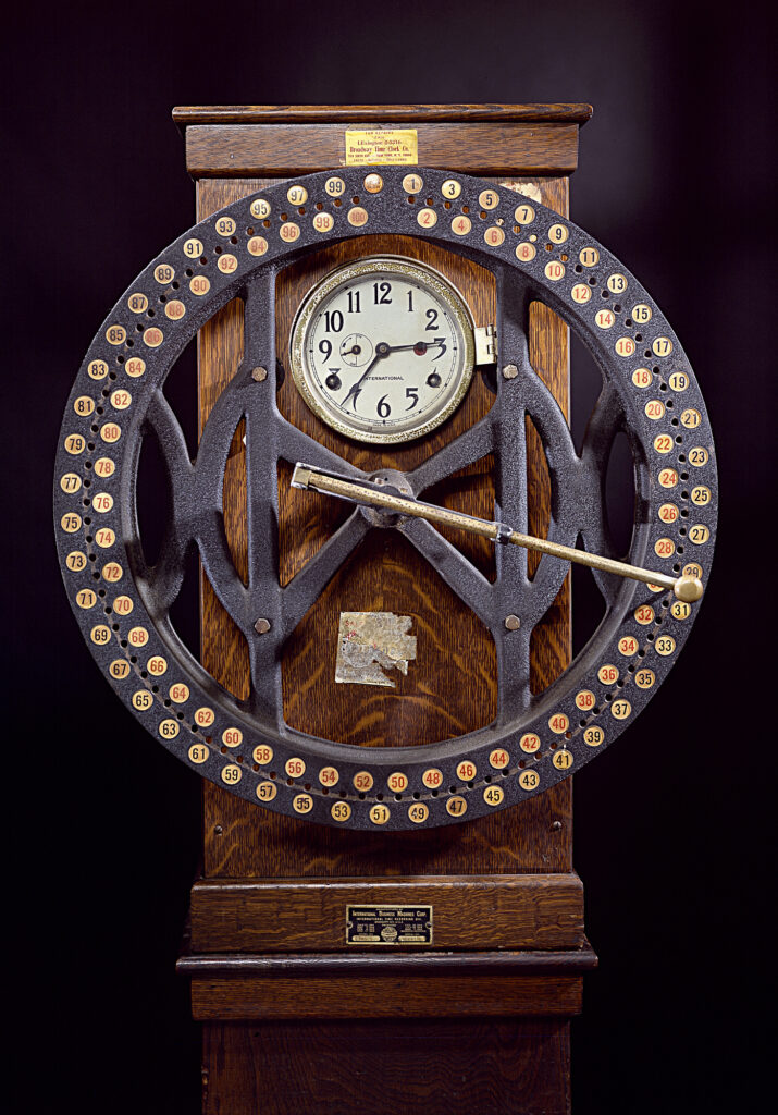 Image description: A closeup of ITR’s dial time recorder; a metal wheel, marked with 100 labelled holes, is attached to a wooden base fitted with a clock. End of alt text.