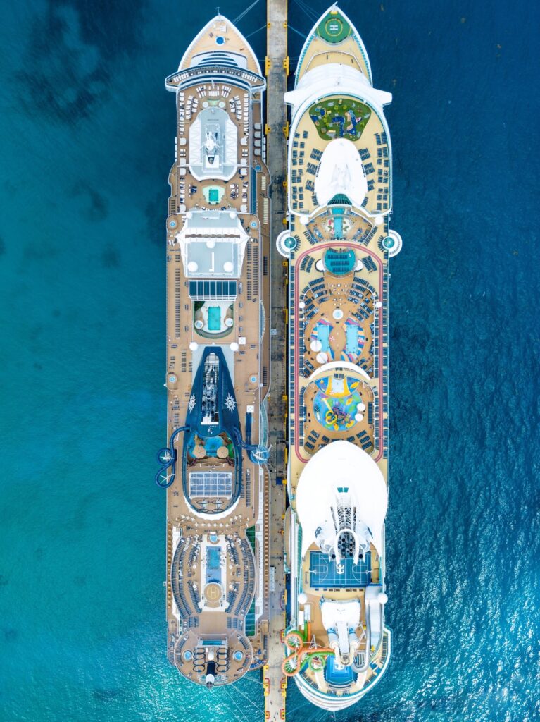 Image description: An aerial view of two large cruise ships docked side by side; both decks have pools, sun loungers and sports facilities. End of alt text.