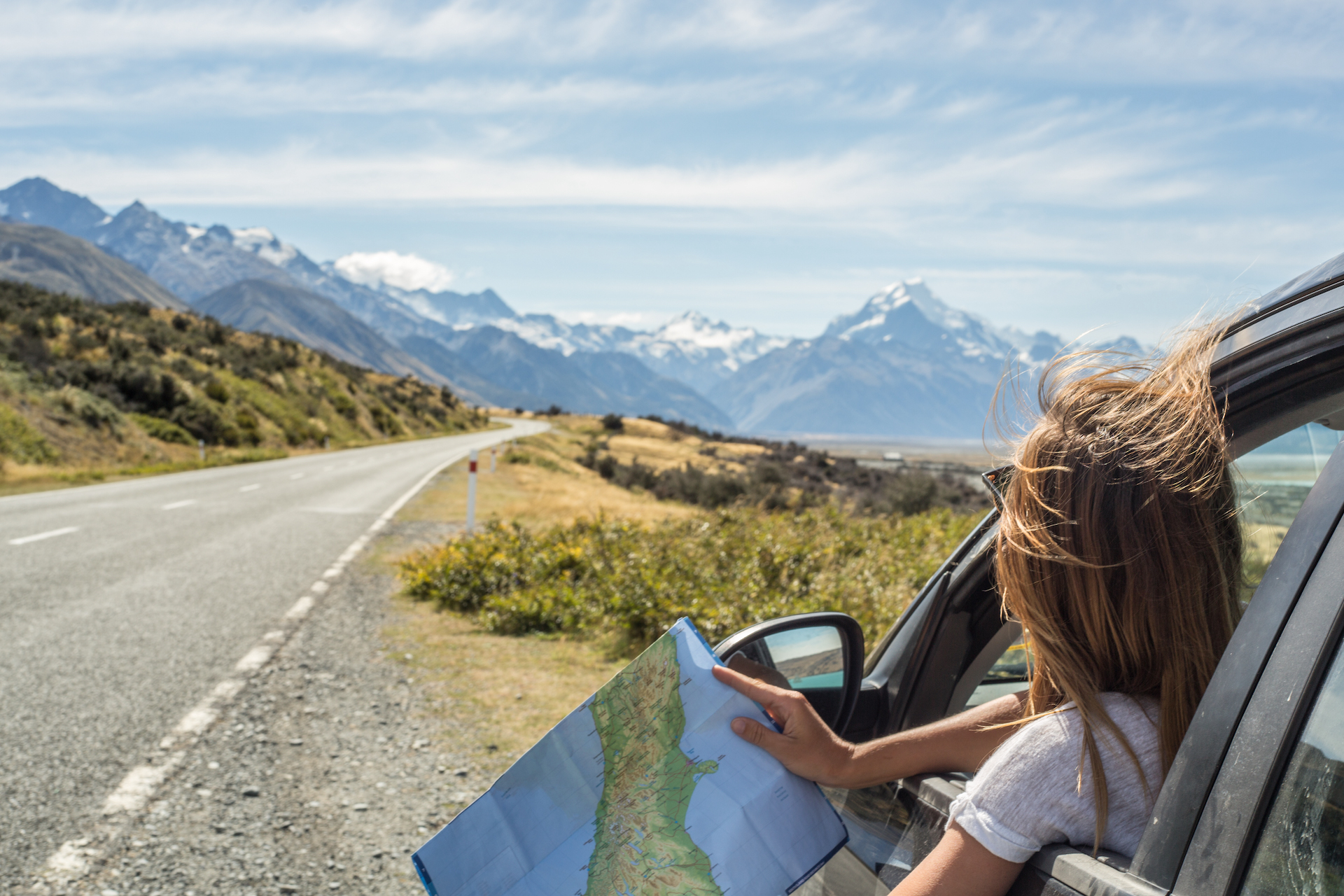 Image description: A person looks out from their car window, with a map of New Zealand in hand, at the view of distant, snow-capped mountains; the car is parked in a lay-by. End of alt text.