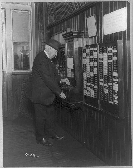 Image description: Thomas Edison punches his time card at his workshop; he is surrounded by a wall filled with time card slots. End of alt text.