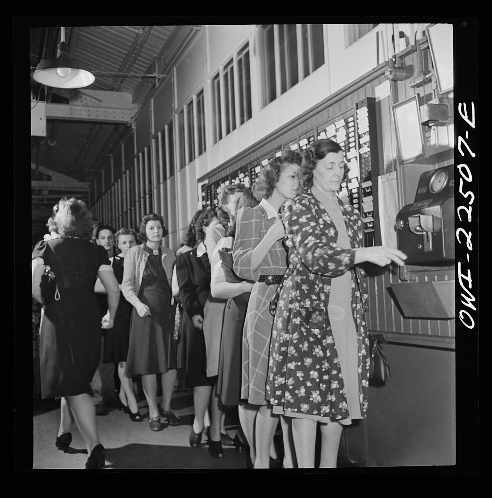 Image description: A vintage photograph of workers lining up to clock into a factory using time cards.  End of alt text.