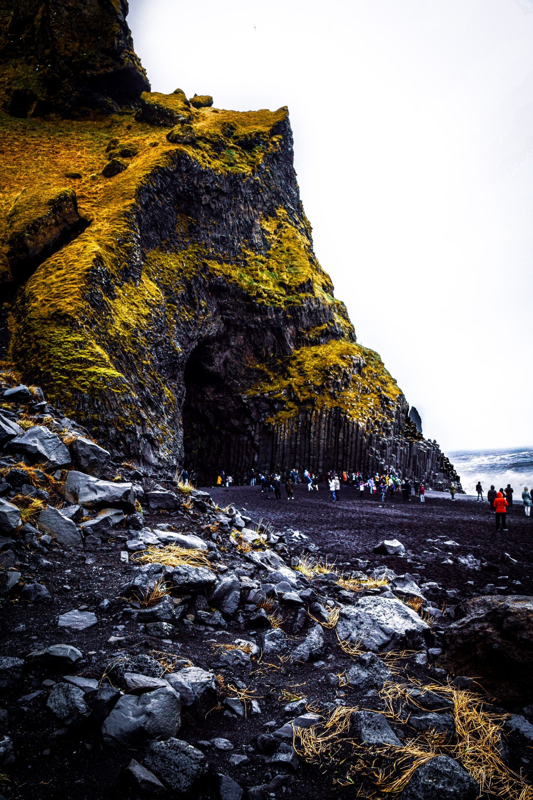 Image description: A black-sand beach, filled with tourists taking photographs, leads to a cave beneath a large, moss-covered cliff in Iceland. End of alt text. 