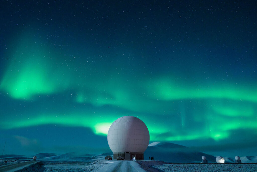 Image description: A polar weather station in Norway; the night sky is illuminated by the green glow of the Northern Lights. End of alt text.