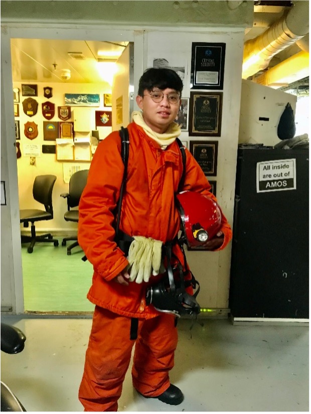 Image description: A full-body portrait of Jewell Lorejo, uniformed all in orange, aboard a ship. End of alt text.