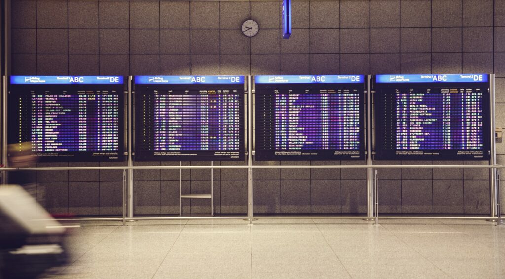 Image description: Four airport departure boards inside a tiled airport concourse. End of alt text.